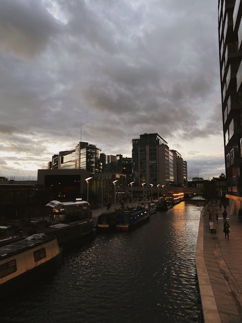 The image depicts a canal scene near Regent's Canal in London during late afternoon or early evening, with a dramatic cloudy sky overhead. On the right side, a modern building with dark glass windows 