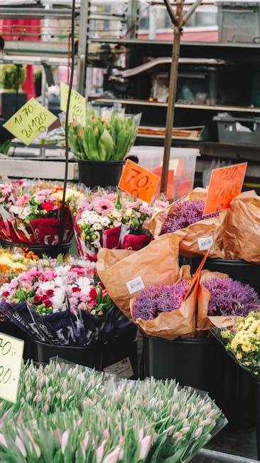 A vibrant display of freshly cut flowers arranged in black plastic buckets at an outdoor market stall. Prominent in the foreground are pale pink tulips with tightly closed buds and green stems, laid o