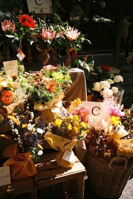 A vibrant display of floral arrangements featuring a variety of fresh flowers, including pink proteas with spiky petals, bright red and orange blossoms, and delicate white and yellow blooms. The bouqu