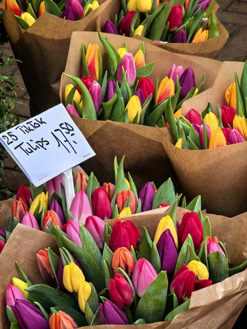 A collection of fresh tulip bouquets with vibrant pink, yellow, orange, and purple blooms, each wrapped in brown kraft paper. The tulips are arranged in a dense, natural style with green leaves and st
