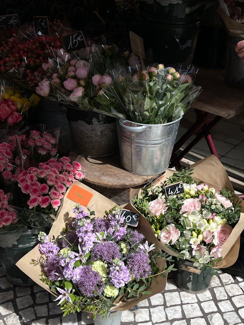 A display of fresh flower bouquets arranged in rustic containers and wrapped in brown craft paper, featuring a variety of blooms including pink roses, purple alliums, and small pink carnations. The bo