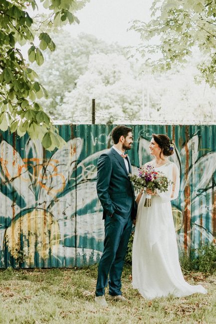 A bride and groom stand outdoors in front of a weathered wooden fence adorned with colorful graffiti, surrounded by green foliage and trees. The bride, holding a floral bouquet, wears a white dress wi
