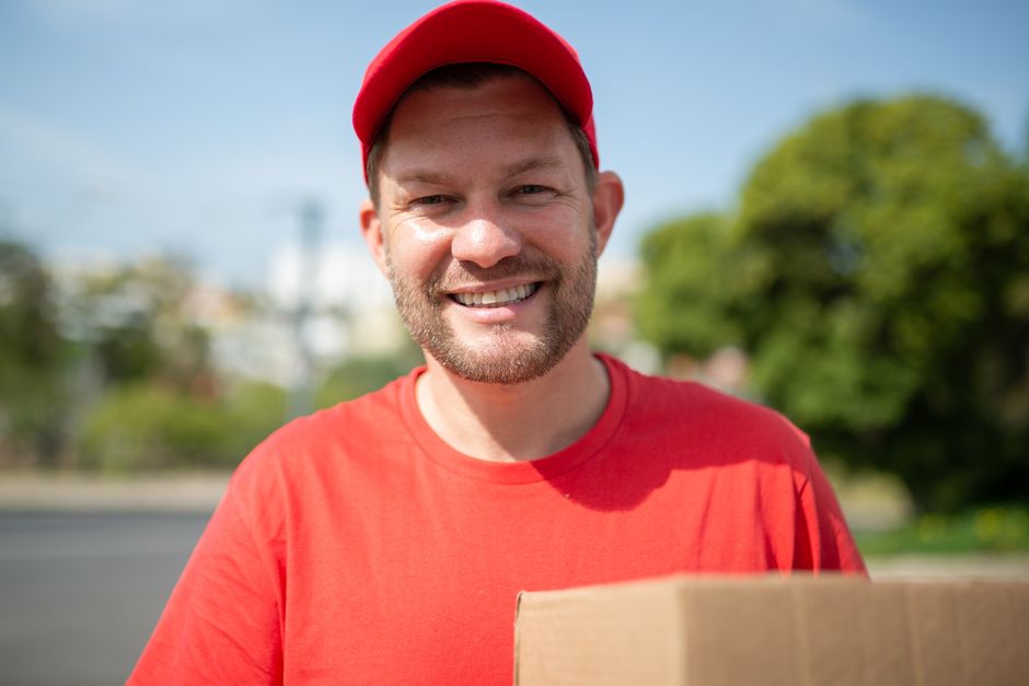 A smiling male flower delivery courier from floristhaggerston.co.uk stands outdoors on a sunny day, wearing a red cap and matching red t-shirt. He holds a cardboard box with both hands, likely contain