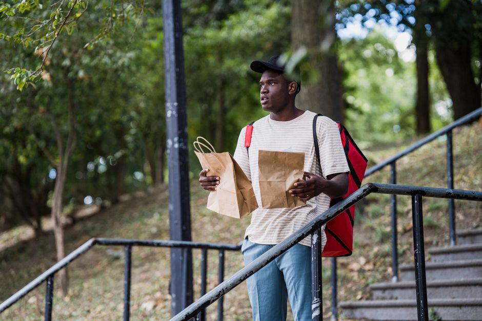 A young man with dark skin and wearing a black cap, a beige striped t-shirt, and light blue pants, is standing outdoors on a staircase surrounded by green trees. He is holding two brown paper bags, on
